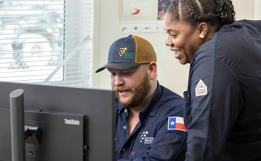 This image features two people interacting with technology. In the foreground, there is an adult male wearing a cap. To his right, there is another person who appears to be female; she is smiling. Both individuals are looking directly at the screen in front of them. They seem to be in an office space, as indicated by the setting and the presence of professional attire