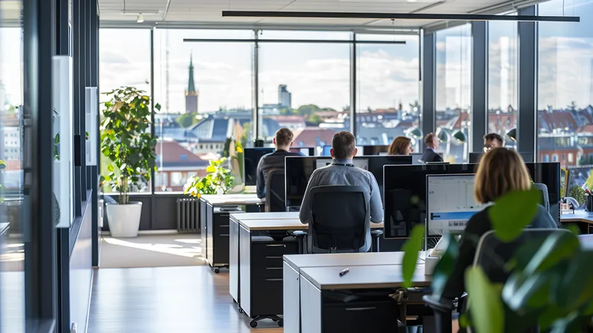The image displays an interior office space with multiple desks and computer workstations. There are people seated at the desks, indicating a shared workspace environment.