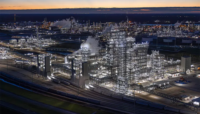Photograph of an industrial complex at night, with a plant that resembles a chemical factory in the center, visible smoke stacks, low-rise buildings, and dark sky.