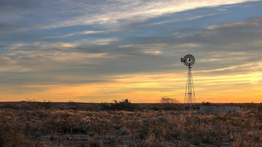 The image shows a serene landscape at sunset. The sky has long, streaky clouds that blend towards the darker parts of the frame, with the warm colors of the sun partially obscured. To the left side of the photo is a silhouette of a windmill against the sky, which stands out as the focal point in the lower part of the image. In front of the windmill and set against a flat horizon, there's an expanse that looks like a desert with no visible vegetation. The field is bordered by an area of bushes or shr