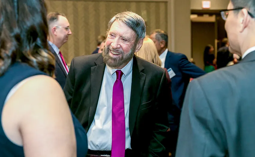 The image shows a person standing among a group of people. This person appears to be engaged in conversation, likely at an event such as a conference or seminar. The individual is wearing a suit with a pink tie and has glasses, suggesting they might be older or middle-aged.