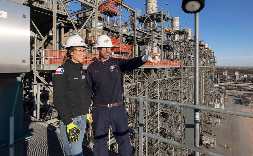 This image features two individuals, one holding a camera and pointing at something off-camera. They are standing on an elevated platform with industrial equipment around them. The environment suggests that they might be working in an oil facility or similar industrial site.
