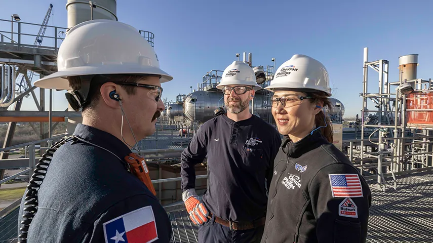 The image shows a group of individuals at an industrial or industrial-like site. They are engaged in a conversation with each other, and one individual is wearing a hard hat, which suggests the presence of ongoing construction or maintenance work. In the background, there are large pipes or conduits which might be related to oil and gas operations due to their size.