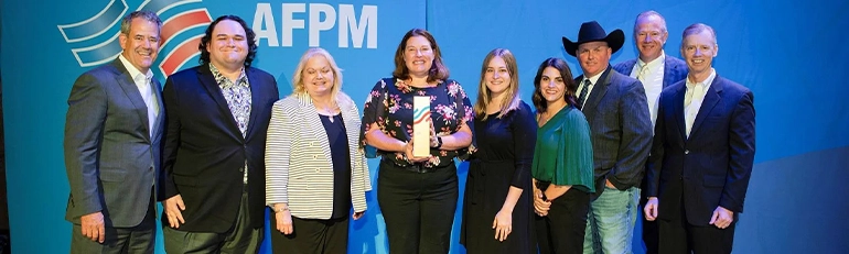 This is an image of a group of individuals posing for a photograph on what appears to be a stage, with a backdrop containing the text "AFPM" and some graphical elements that suggest a professional or organizational context. The people in the photo are dressed formally and are smiling towards the camera. There's a prominent person at the center holding an award or a certificate, which seems to be the focus of their gathering.