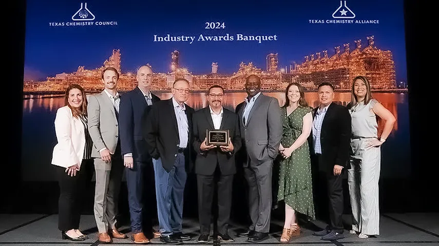 This is a photograph of a group of individuals standing in front of what seems to be an award or recognition plaque. The setting is an indoor event, as indicated by the stage backdrop and the presence of lighting rigs. The plaque is titled "IDRA Industry Pioneers Banquet 2024".