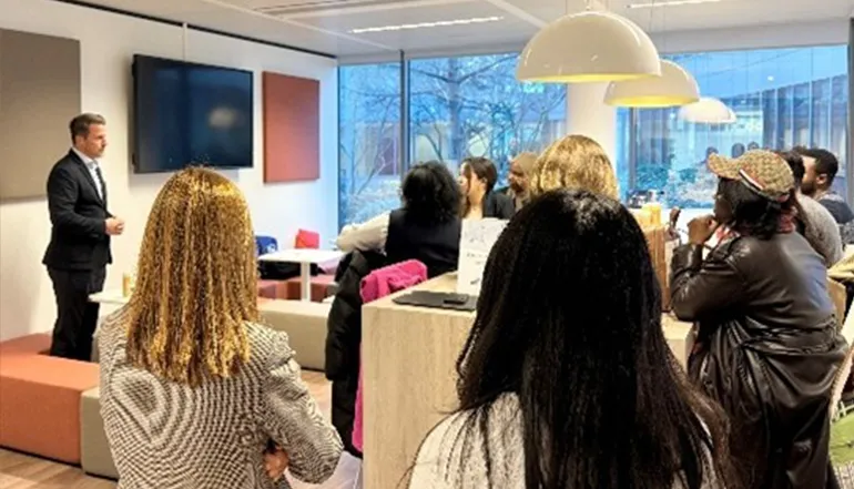 This is an image that appears to show an indoor setting where a person, likely leading or moderating the meeting, stands standing front of a small group of participants seated around tables. It looks like they are engaged in a presentation or discussion, possibly during a business meeting or workshop. The setting suggests a professional environment, and there's a display screen showing a slide presentation that matches the content on the speaker's laptop. Everyone seems focused on the presenter, and the atmosphere appears to be attentive and interactive.