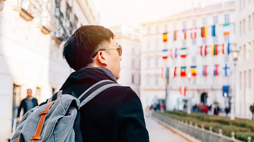 The image shows a man standing with his back to the camera. He appears to be walking down an urban street lined with shops and buildings.