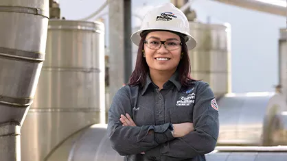 The image shows a person standing in front of an industrial setting, possibly at a refinery or similar facility. This individual seems to be in work clothes, commonly associated with safety attire for industrial environments.