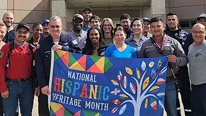The image depicts a group of people posing in front of a sign. It seems like an event or celebration related to "National Hispanic Heritage Month," as indicated by the text on the sign, which includes colors and images that likely represent aspects of Hispanic tradition and culture. There's also a tree with rainbow colors and shapes, symbolizing diversity or support for LGBTQ+ rights. The people in the photo are dressed in various styles of clothing, indicating a casual gathering open to the public.