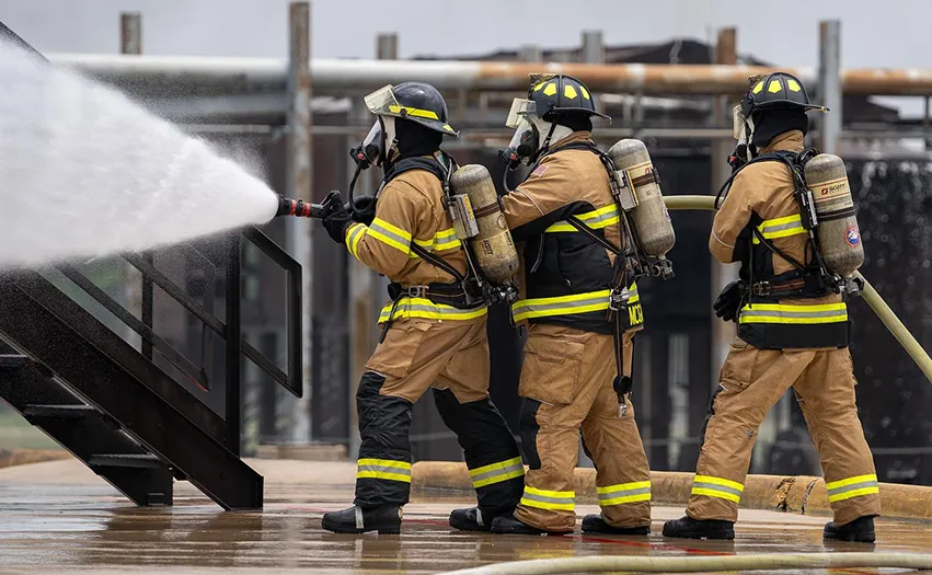 This image shows three firefighters in full gear, working with water hoses. The firefighter in the center appears to be using a hose and nozzle while another two individuals stand nearby, possibly ready to assist or connected to additional suppres equipment not visibly in the picture. They are outdoors during what seems like normal daylight. There is a structure that resembles scaffolding or temporary construction platform behind them. The firefighting gear indicates they are either actively engaged in firefighting operations or carrying out a training exercise for such situations.