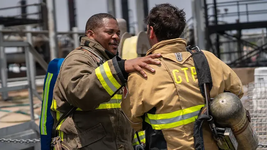 The image depicts two individuals in what appears to be firefighting gear, engaging in a seemingly friendly interaction.