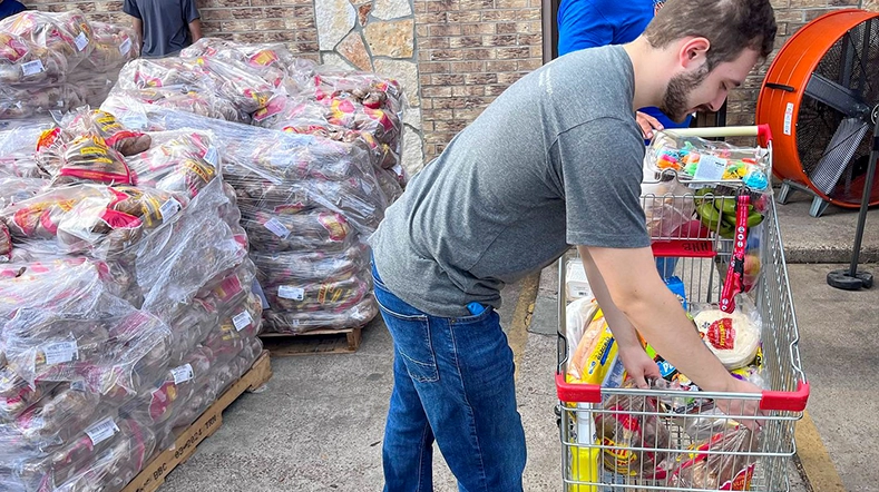 The individual is wearing a gray t-shirt and denim jeans, looking to their left while facing away from the camera. There are various items on the pallets, including cases of water bottles, some products visible among them, suggesting this might be an event or community relief effort. No clear face features are visible in the image, making the identity of the person unidentifiable. The setting seems to be outdoors with a brick wall and what looks like a cardboard
