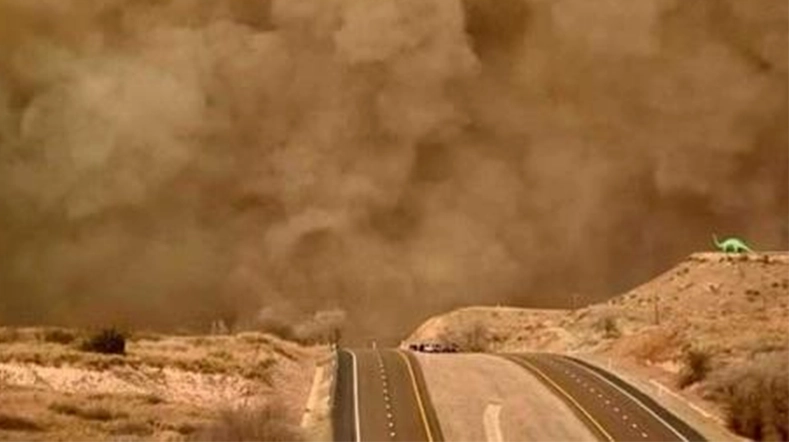 This image shows a wide view of a highway with road signs, surrounded by the foreground dominated by blowing sand. In the background, there are brownish clouds and haze, suggesting that this area is experiencing heavy dust and possibly a significant storm or windstorm. 