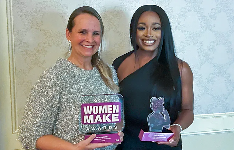 The image shows two individuals at a function, holding plaques. They are standing side by side with smiling expressions on their faces. One person is standing slightly behind the other. On the left, there's a woman holding an award titled "WOMEN MAKE Awards."