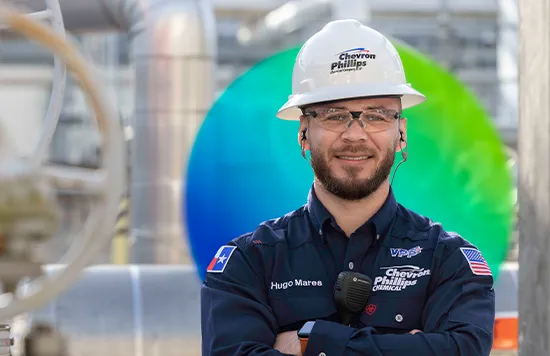 The image shows an individual wearing a hard hat and holding what appears to be a digital device, possibly for communication or data analysis. The person is posing in front of industrial equipment, indicative of an oil or gas plant environment. In the background, there are tanks with various labels and logos, suggesting a process industrial setting.