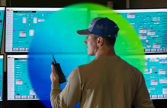 The image shows a person standing in front of numerous TV monitors displaying various graphics, charts, and tables that appear to be related to data analysis, system monitoring, or control room operations. The individual is holding a device resembling a police scanner to their ear, possibly listening to radio communications. Atop the monitors is what looks like a security badge, suggesting restricted access to this area. 