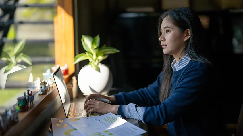 A woman working on a laptop, looking out the window in front of her with a thoughtful face expression. Outside it's green and sunny. (Photo)