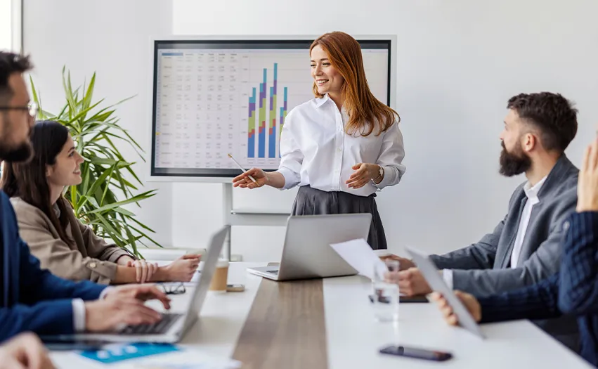 A woman leading a meeting in an office with some charts on a big screen in the background (Photo)