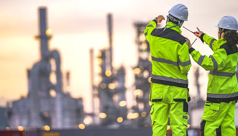Two people with safety vests, helmets, and walkie talkies pointing at something on an industrial site (Photo)