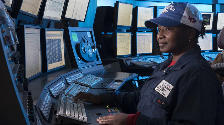A person wearing a uniform and cap working at a control panel with multiple monitors and keyboards. (Photo)