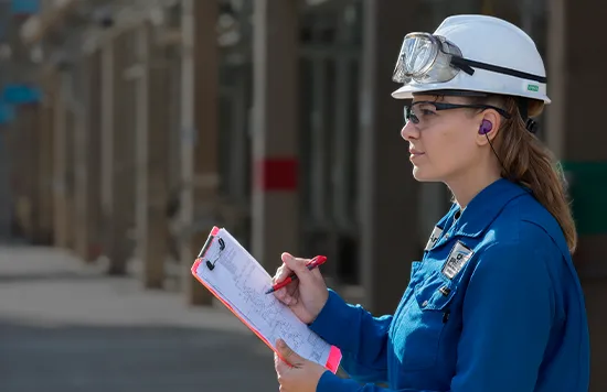 A woman with a uniform, safety googles, and a helmet at a production site. She's holding a clipboard and a pen (Photo)